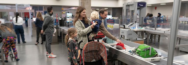 A family at an airport security screening checkpoint; a woman is holding a toddler and a stroller is in front of them. On the left of the woman is a young girl and on the right is a man putting a ball cap in a bin for X-ray screening.