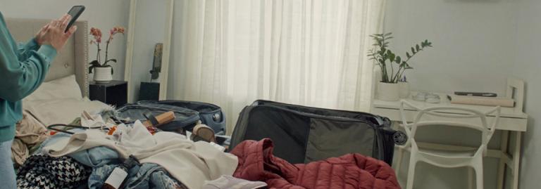 Person packing a suitcase on a bed cluttered with clothes and travel essentials in a bright bedroom.