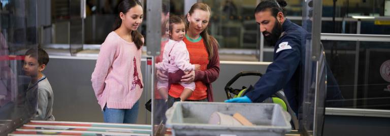 Une passagère tenant un enfant en bas âge reçoit l'aide d'un agent de contrôle pour retirer ses effets personnels.