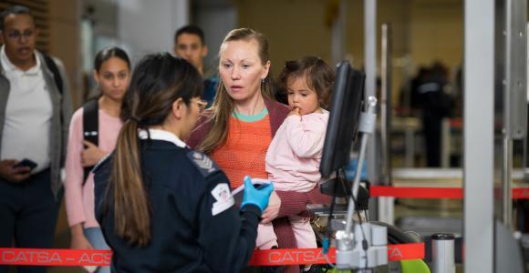 A female passenger holding a toddler is handing her electronic boarding pass to a female screening officer.