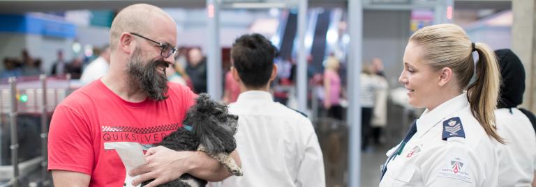 A passenger going through airport security screening while holding their dog and a paper boarding pass while a screening officer watches.