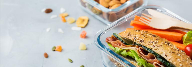 A lunch container on a countertop is filled with nuts, seeds and dried fruits. Another container beside it has a deli sandwich, sliced vegetables and wooden cutlery.