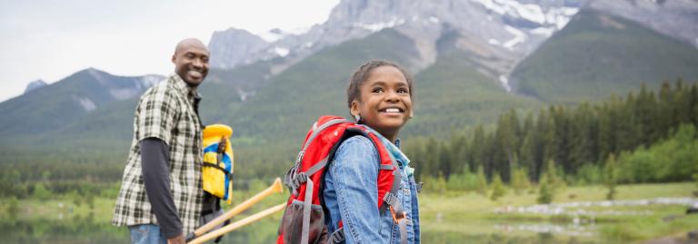 Un adulte et un enfant, équipés pour faire du canot, sourient devant un lac avec des montagnes en arrière-plan.