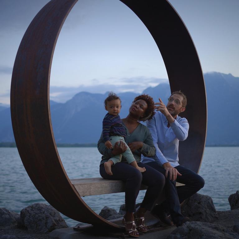 Famille assise sur une sculpture circulaire au bord d'un lac, avec vue sur le lac et les montagnes.